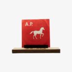 Red square card with a white running horse silhouette and the letters "A.P." mounted on a wooden base with a black support piece behind it