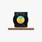 Black vinyl record mounted on a black display block set on a wooden base, with a colorful label at the center of the record.