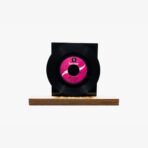 Black vinyl record mounted on a black square pedestal atop a wooden base with a pink label.
