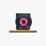 A black vinyl record on a wooden stand against a light background, with a pink label in the center and a dark square backing behind it.
