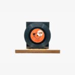 Black vinyl record mounted on a square black stand placed on a wooden platform, with an orange label visible at the center of the record.