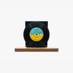 Black vinyl record mounted on a wooden display base with a blue-yellow center label, set against a white background.