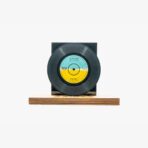 Black vinyl record mounted on a square black stand placed on a wooden base against a white background, museum-style display piece.