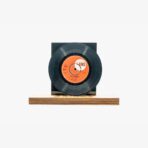 Black vinyl record mounted on a dark block atop a wooden base, centered on a white background, like a sculpture or display piece with an orange label in the center