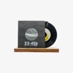 Black vinyl record partially slid out of a dark gray sleeve with a circular window, resting on a wooden base; the sleeve reads '33:45'.