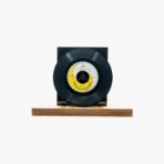 Black vinyl record mounted on a wooden pedestal against a white background, showing a yellow-label center.