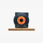 Black vinyl record mounted on a small black cube atop a wooden base against a white background, with an orange label at the center.