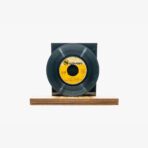Black vinyl record mounted on a wooden base with a dark backdrop and a yellow label at the center.