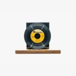 Black vinyl record with yellow label on a black stand atop a wooden base, displayed like a sculpture or display piece.