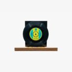Black vinyl record mounted on a wooden platform with a blue‑yellow label, against a white background for display purposes.