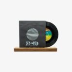 Black vinyl record partially pulled from a dark sleeve with a circular window, showing '33:45' on the cover, resting on a wooden base.