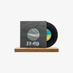 Black vinyl record partly inside a dark sleeve with a circular window, displayed on a wooden stand, '33:45' printed on the sleeve edges the front.