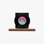 2301450048D | 3345 Records Black vinyl record mounted on a wooden pedestal against a white background, with a pink-and-turquoise label at the center of the disc.