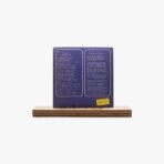 Purple square book/plate with two white outlined text panels on a wooden display base, small yellow sticker in bottom-right corner