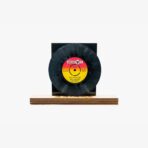 Vinyl record mounted on a black display with a yellow–red label centered, resting on a wooden base.