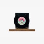 Black vinyl record mounted on a wooden platform, label facing outward, against a white background on display.