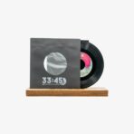 Black vinyl record partially pulled from a gray sleeve with a circular window, sleeve shows '33:45' and sits on a wooden base.