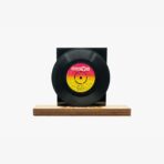 Black vinyl record mounted on a square black stand resting on a wooden base, label visible with yellow–pink gradient design and text