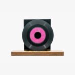 Black vinyl record with a pink center label mounted on a wooden display base.