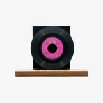 Black vinyl record mounted on a dark square display block, with a pink center label, set on a wooden base.
