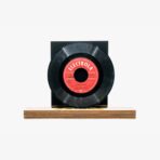 Vinyl record mounted on a black square stand resting on a wooden base with a red label at the center.