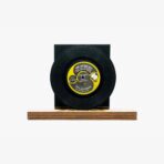 Black vinyl record mounted on a black square stand with a wooden base, showing a yellow label in the center.