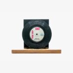 Black vinyl record mounted on a dark rectangular stand atop a wooden base, displayed as art.