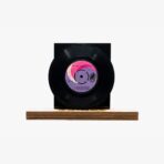 Black vinyl record mounted on a black square stand atop a wooden base, pink label visible in the center