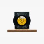 Black vinyl record mounted on a wooden display stand with a yellow label at its center, against a white background.