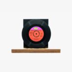 Vinyl record mounted on a black square stand atop a wooden base against a white background, showing an orange label at the center.