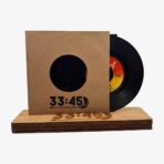 Vinyl record partially pulled from a brown cardboard sleeve with a circular window, resting on a wooden display base; orange-yellow label is visible.
