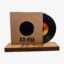 Vinyl record partially pulled from a brown cardboard sleeve with a circular window, resting on a wooden display base; orange-yellow label is visible.
