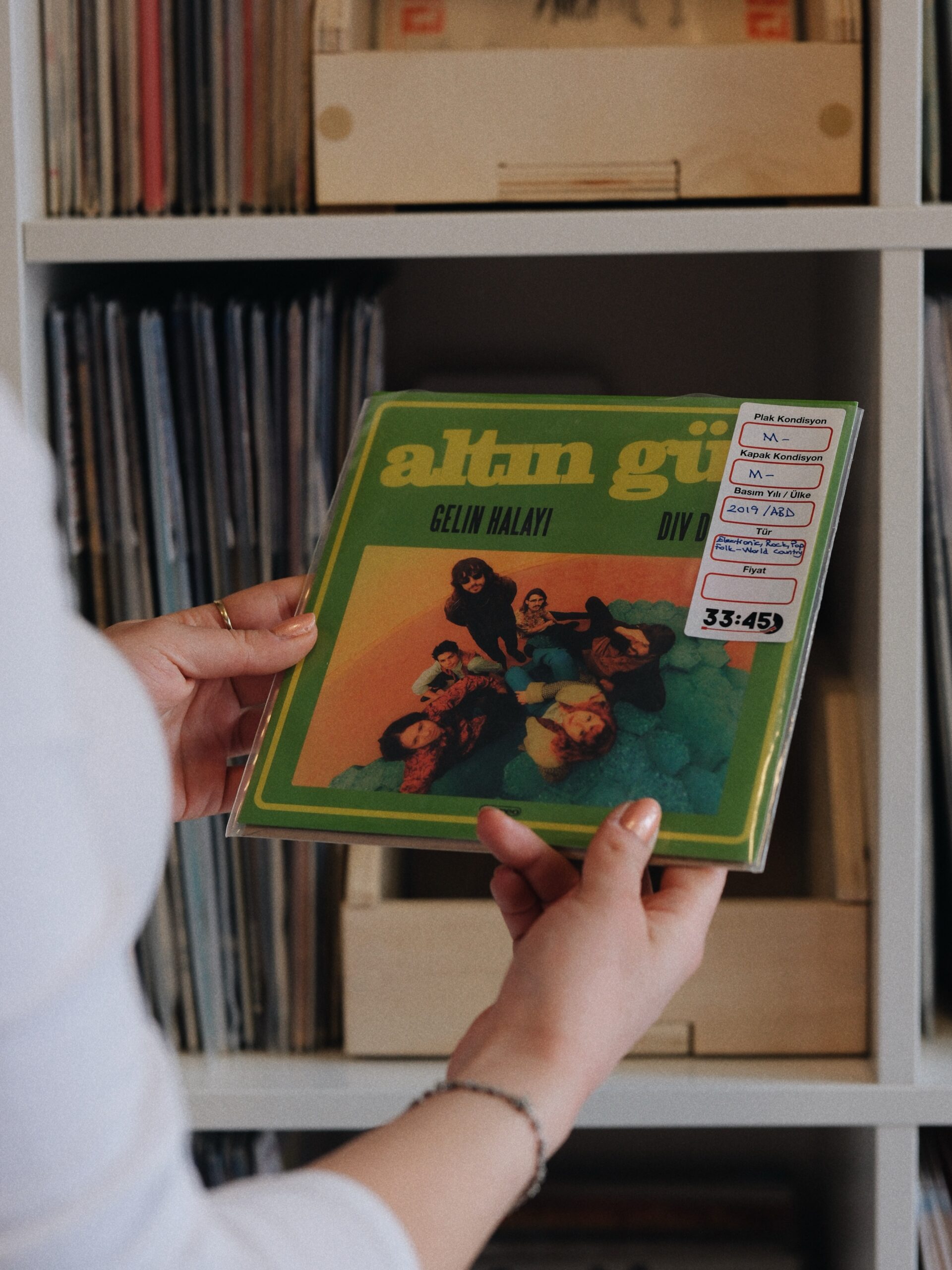 Person holding a green vinyl record in a clear sleeve near a shelf of records.
